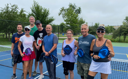Group of people smiling and standing together in front of a pickleball net.